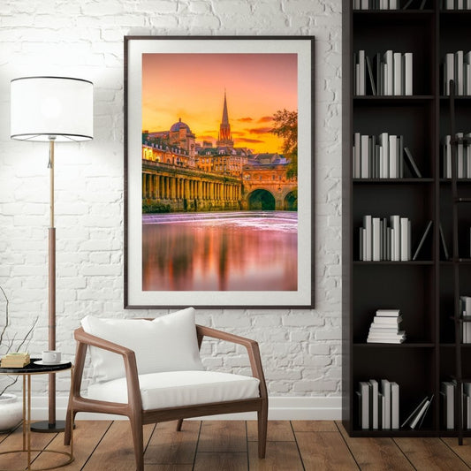 Framed cityscape print on a wall above a wooden chair with a lamp and bookshelf in a room.