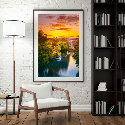 Framed artwork of a sunset over a river with a chair and lamp in a room.