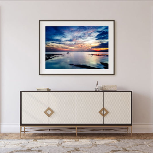 Framed artwork of a sunset over water above a decorative cabinet.