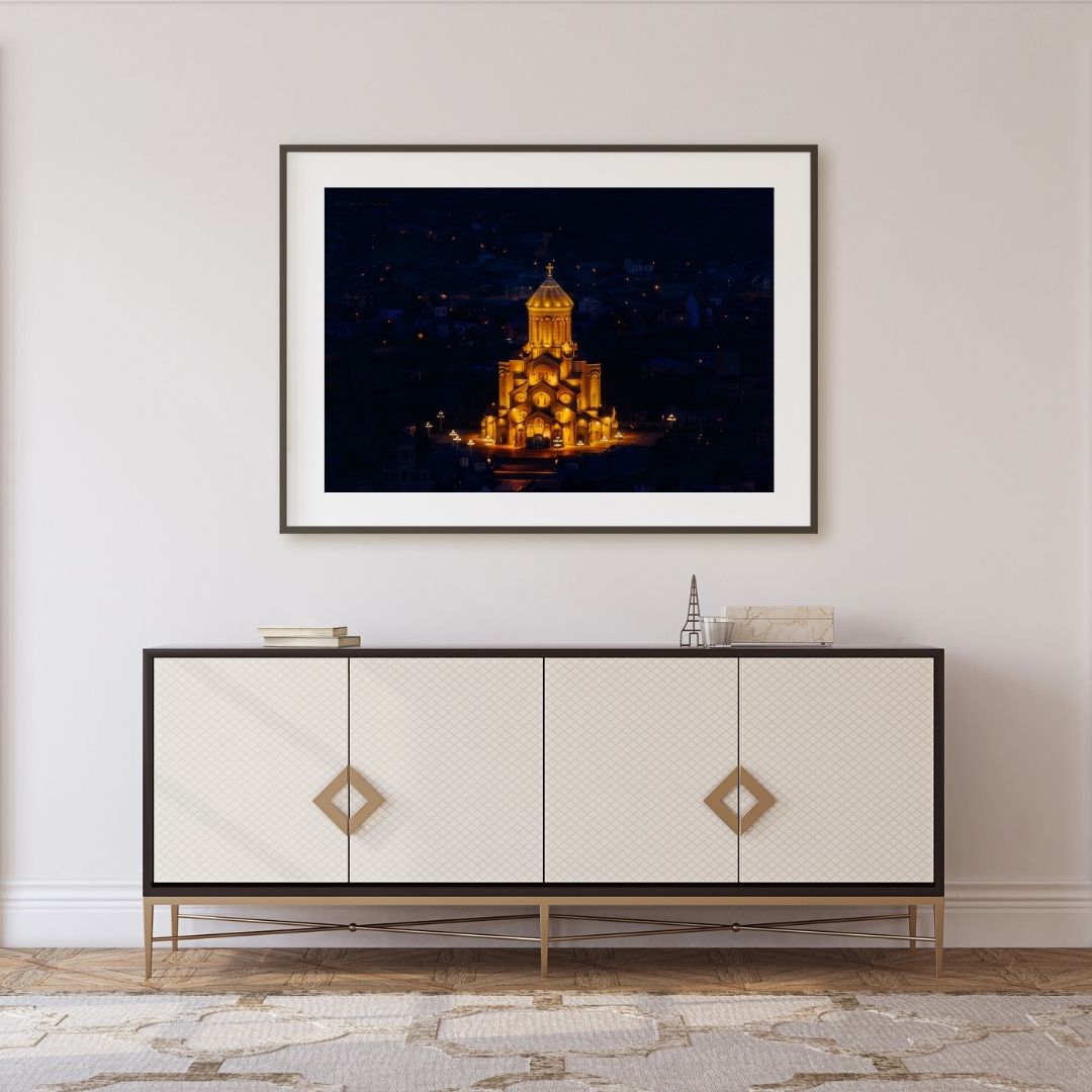 Framed artwork of a illuminated building on a wall above a decorative cabinet.