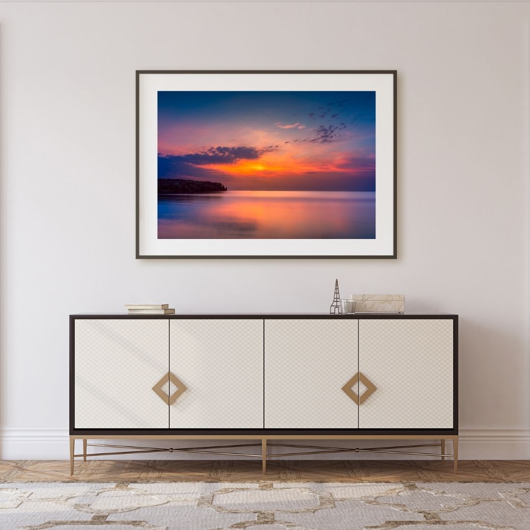 Framed artwork of a sunset over water above a decorative cabinet.