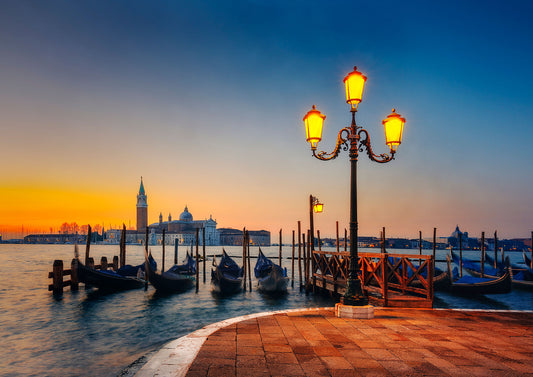 Venetian gondolas moored at sunrise with glowing street lamps and San Giorgio Maggiore in the background