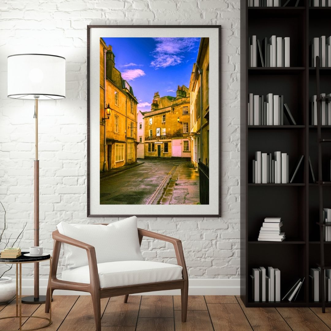 Framed artwork of a street scene on a white brick wall above a wooden chair.