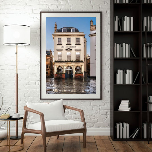 Framed photograph of a building on a wall in a room with a chair, lamp, and bookshelf.