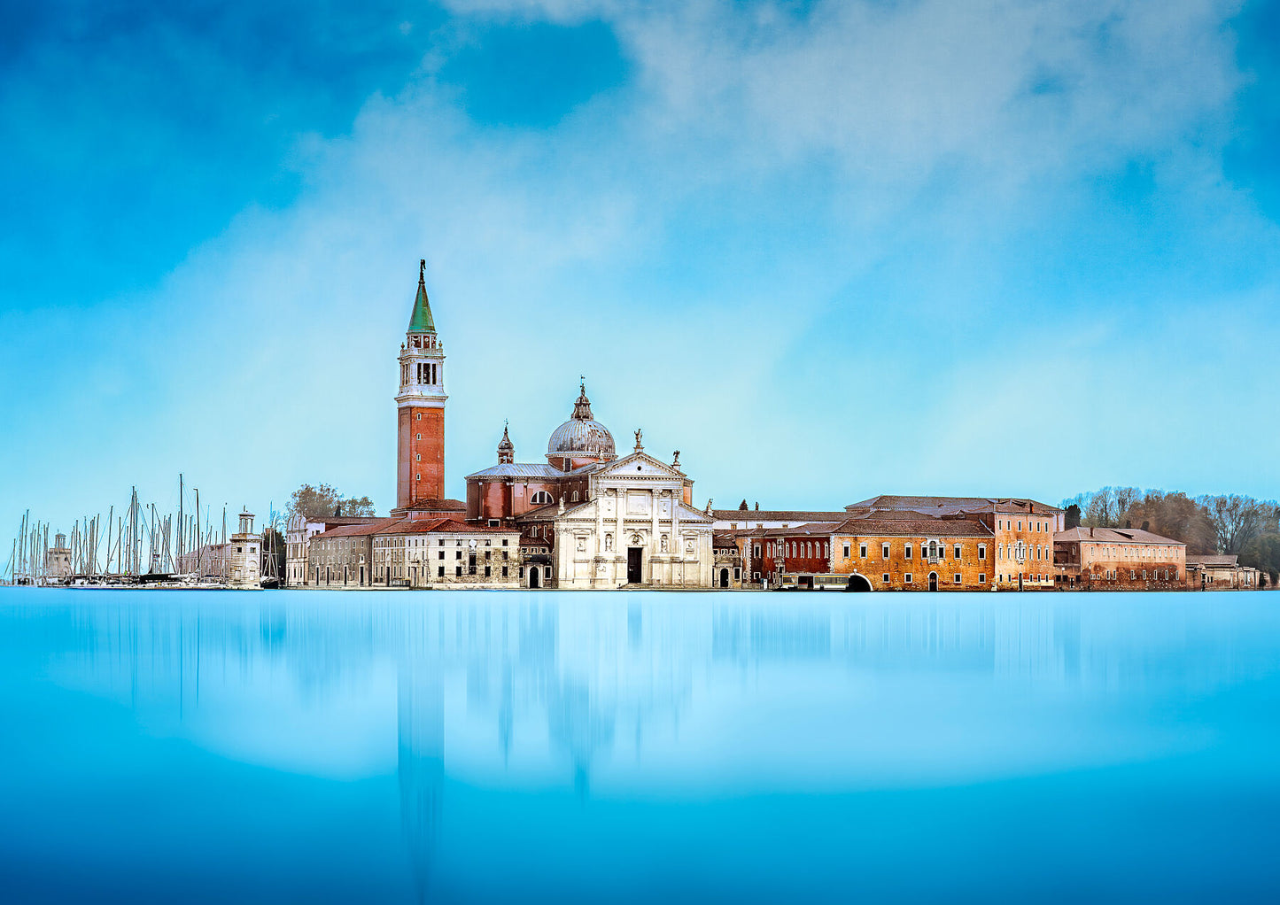 Fine art photograph of San Giorgio Maggiore in Venice reflected in the calm blue waters of the lagoon, with a softly darkened sky and historic architecture.