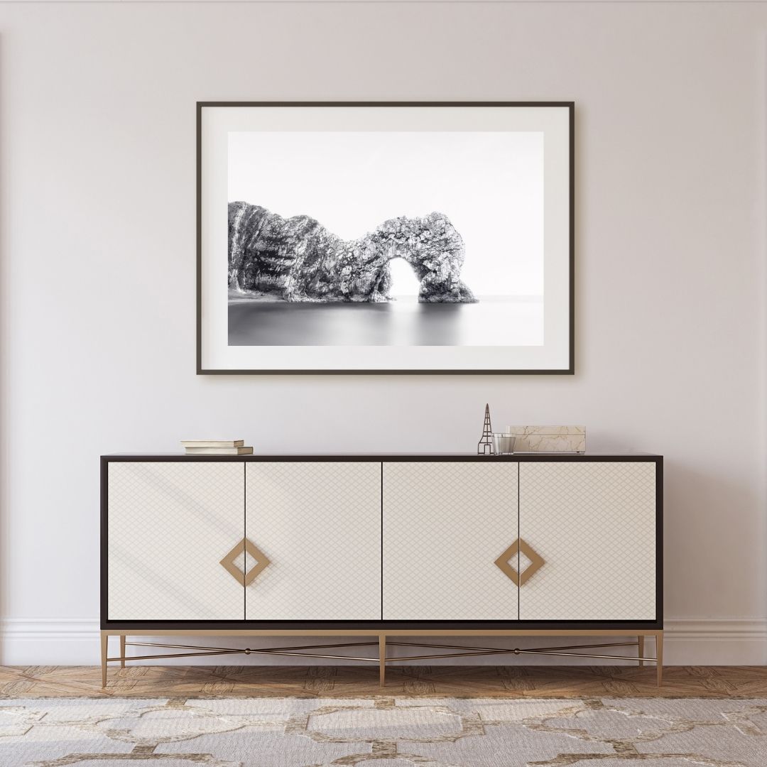 Framed black and white artwork above a decorative cabinet in a room with light-colored walls and flooring.