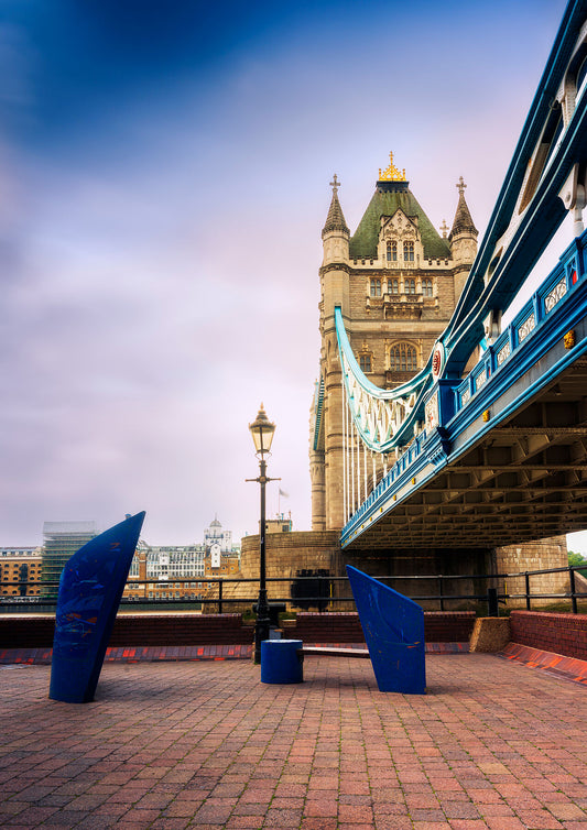 Fine art photo of Tower Bridge in London taken from the riverside walkway, featuring the stone tower, suspension cables, and a modern blue sculpture bench under soft early morning light.