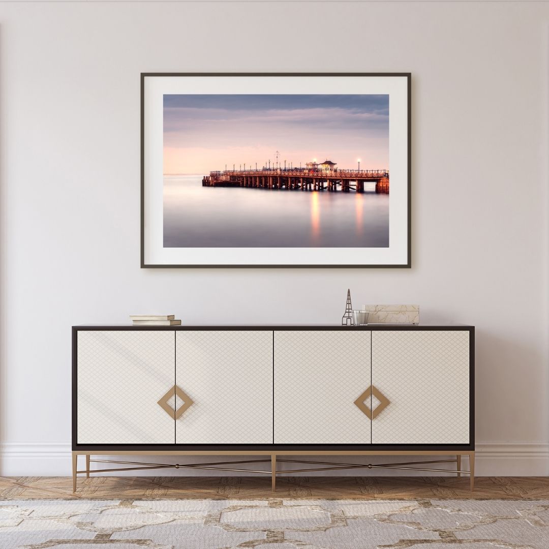 Framed artwork of a pier over water on a wall above a decorative cabinet.