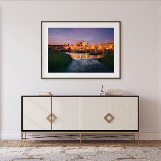 Framed artwork of a sunset over a bridge over water on a wall above a decorative cabinet.