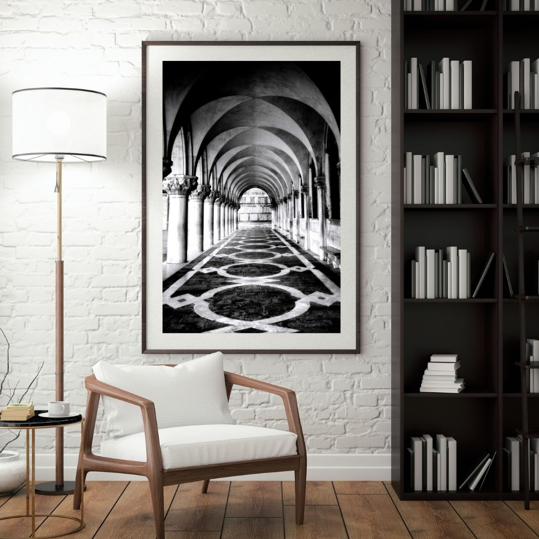 Framed black and white artwork on a brick wall above a wooden chair with a lamp and bookshelf in a room.
