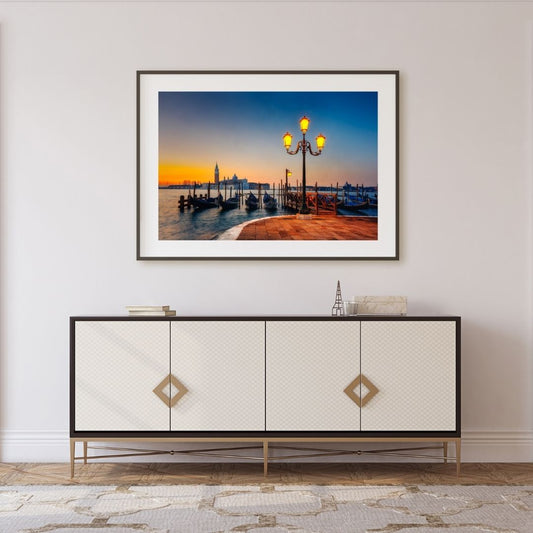 Framed photograph of a sunset over a waterfront scene above a decorative cabinet.
