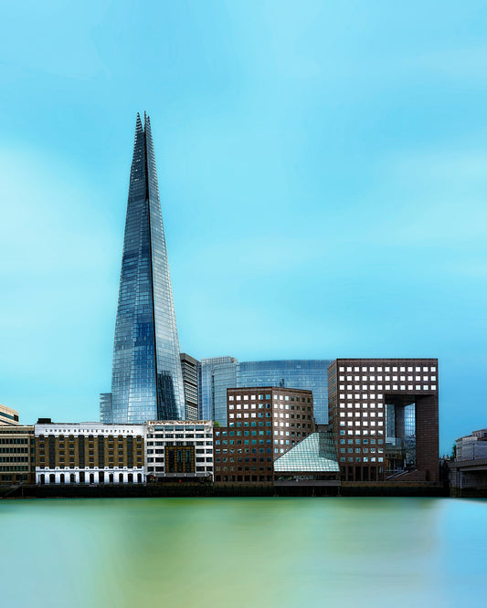 Fine art photograph of The Shard and surrounding Southbank buildings in London with a smooth Thames foreground and pale cyan sky.