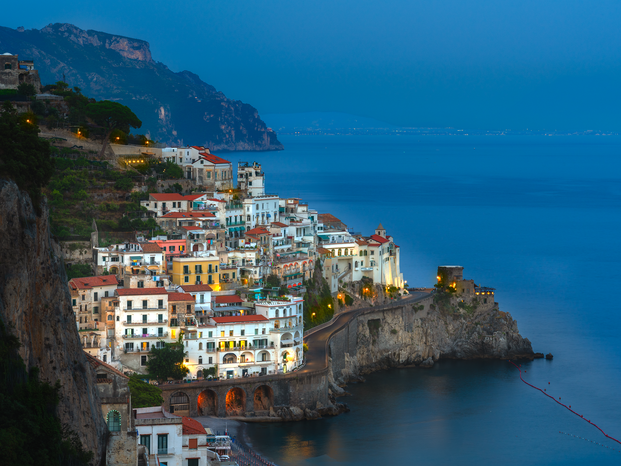 Colour photograph of Amalfi Italy at blue hour showing cliffside buildings warm lights and calm sea under deep blue sky.