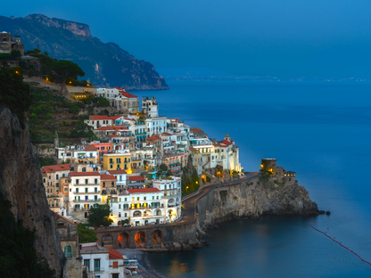 Colour photograph of Amalfi Italy at blue hour showing cliffside buildings warm lights and calm sea under deep blue sky.
