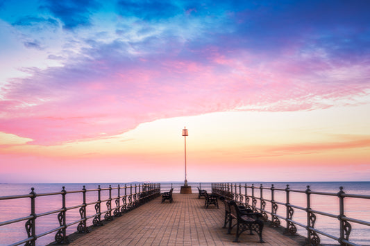 Pastel sunset over Banjo Pier with calm sea, symmetrical railings, and a central lamppost leading into the horizon.
