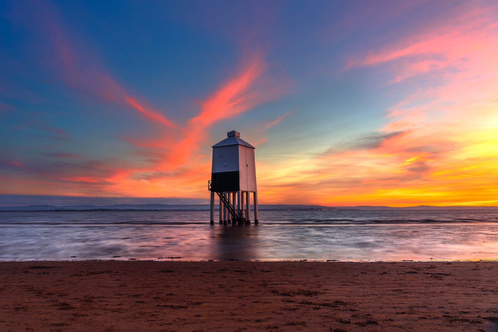 Burnham Lighthouse standing in shallow water at sunset, with warm sky colours, calm sea, and a tranquil coastal atmosphere.