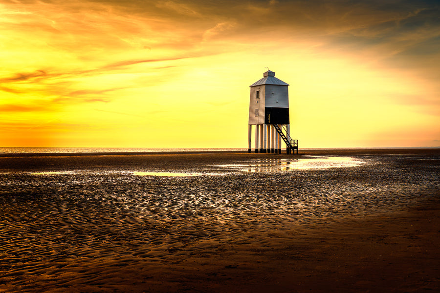 Colour photograph of Burnham-on-Sea lighthouse at sunset with golden sky tidal beach patterns and shallow water reflections.