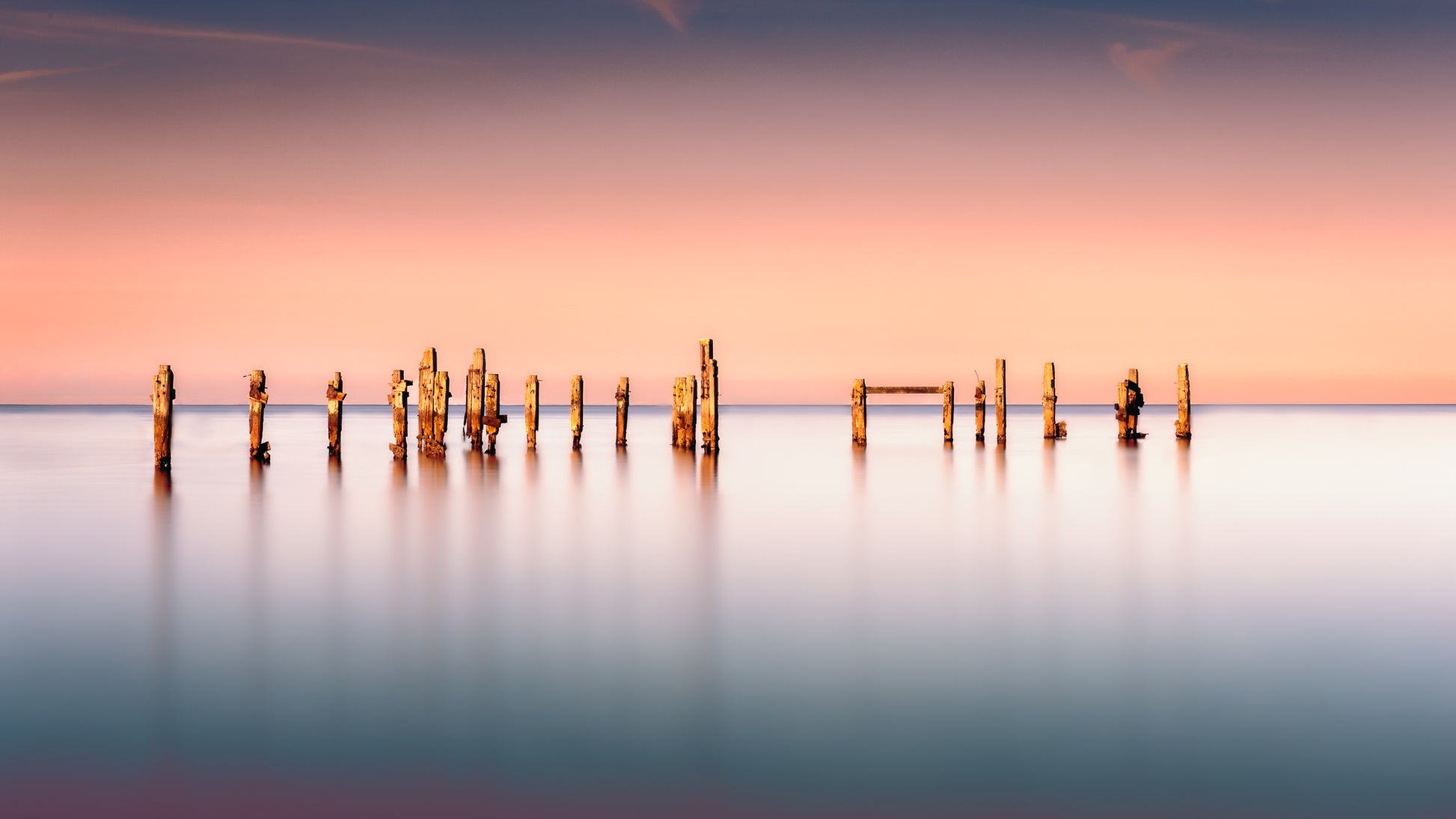 A fine art colour photograph of the old pier at Swanage, England, showing weathered posts in calm water during a warm evening long exposure, created by John FR Wright.