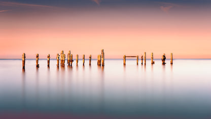 A fine art colour photograph of the old pier at Swanage, England, showing weathered posts in calm water during a warm evening long exposure, created by John FR Wright.