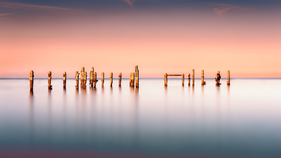 A fine art colour photograph of the old pier at Swanage, England, showing weathered posts in calm water during a warm evening long exposure, created by John FR Wright.