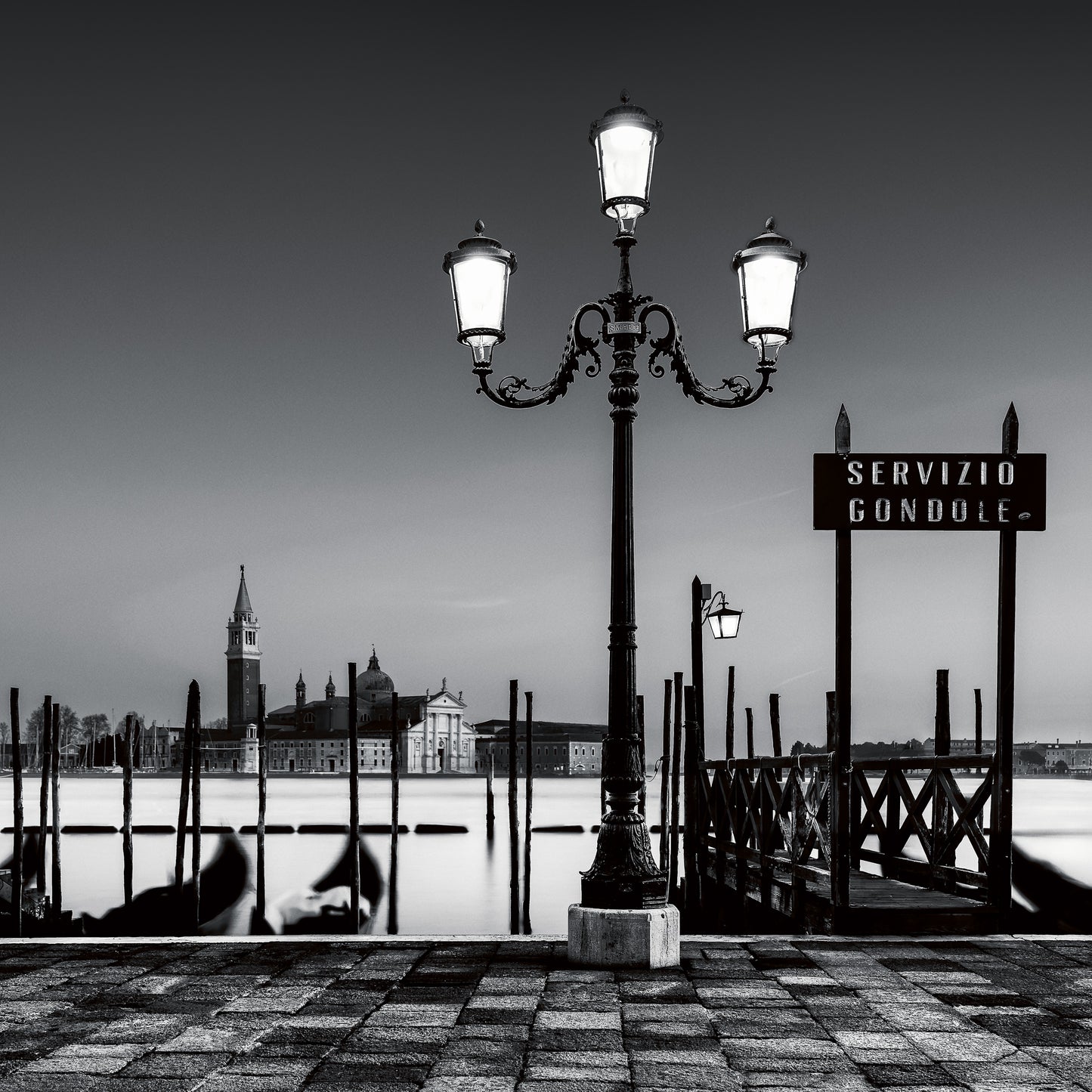 A fine art black and white photograph of a lamppost and gondola service area in Venice, with San Giorgio Maggiore in the background and calm long-exposure water.