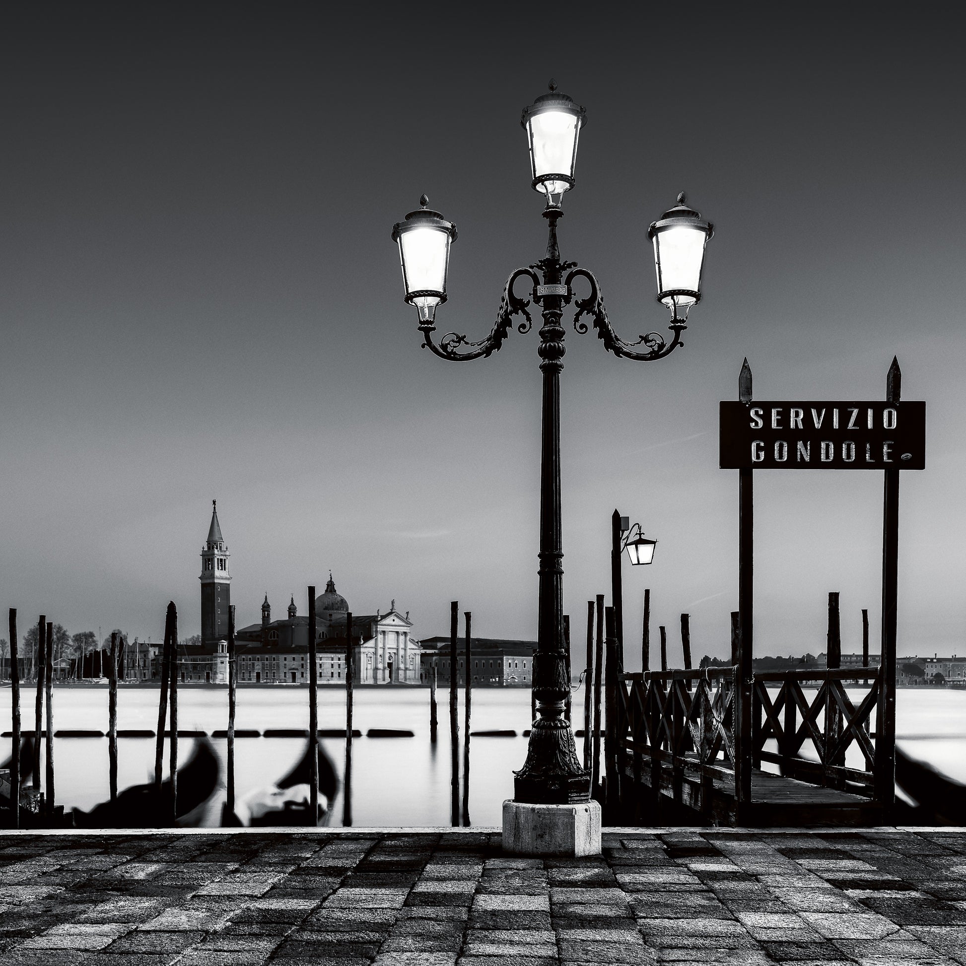 A fine art black and white photograph of a lamppost and gondola service area in Venice, with San Giorgio Maggiore in the background and calm long-exposure water.