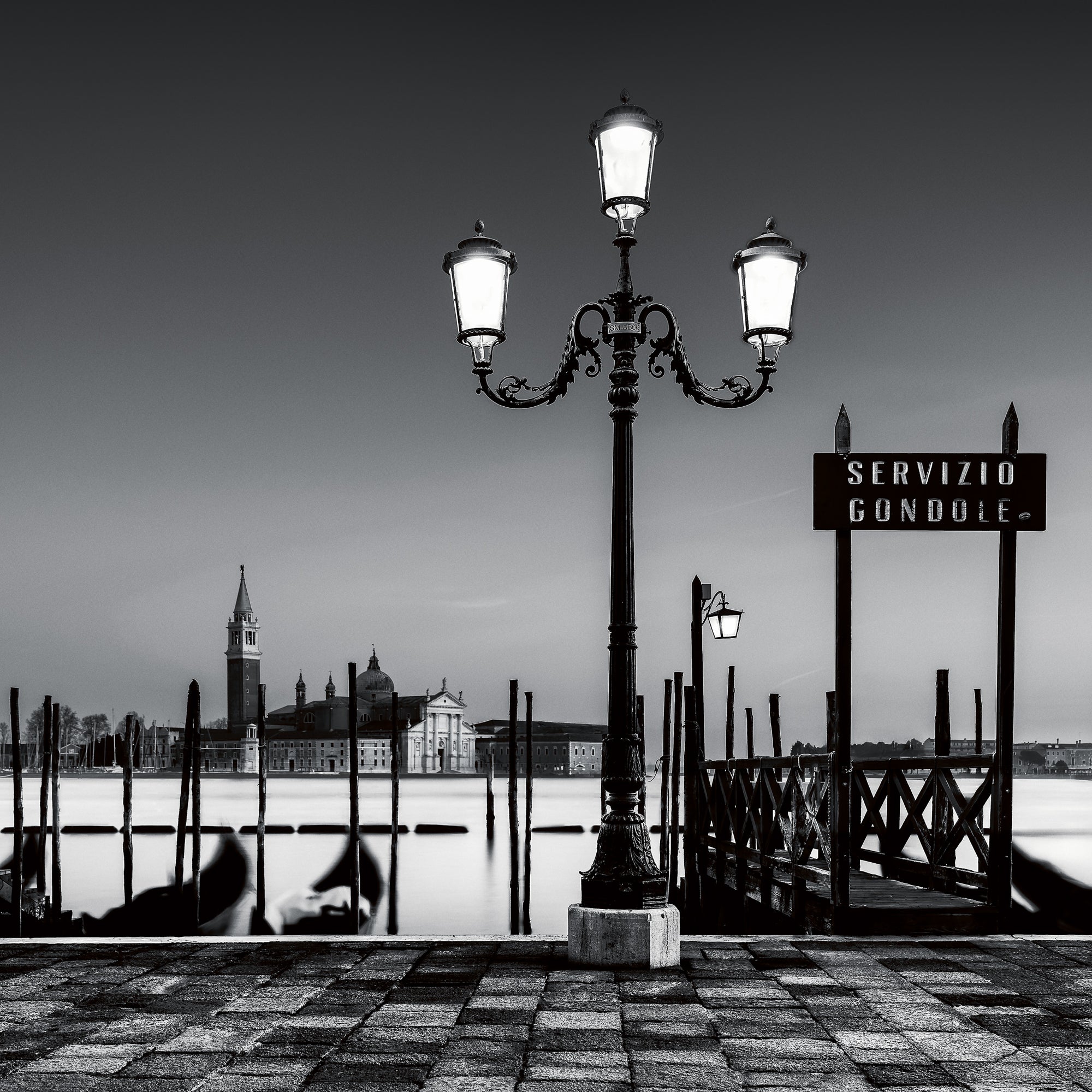 A fine art black and white photograph of a lamppost and gondola service area in Venice, with San Giorgio Maggiore in the background and calm long-exposure water.