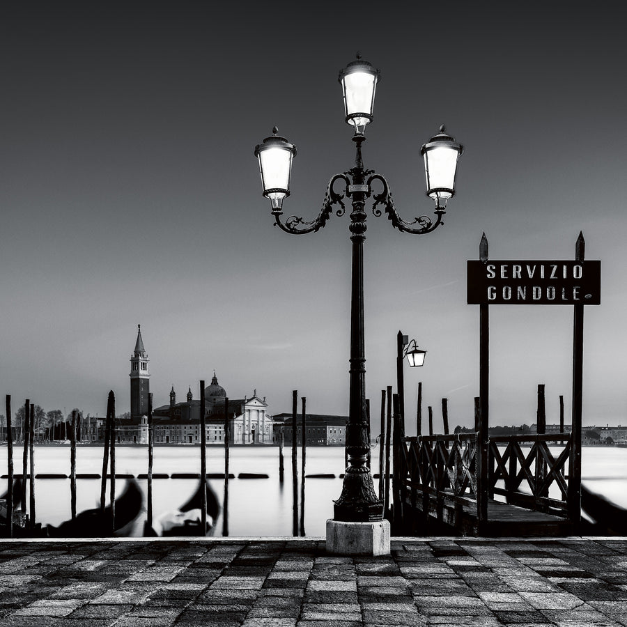 A fine art black and white photograph of a lamppost and gondola service area in Venice, with San Giorgio Maggiore in the background and calm long-exposure water.