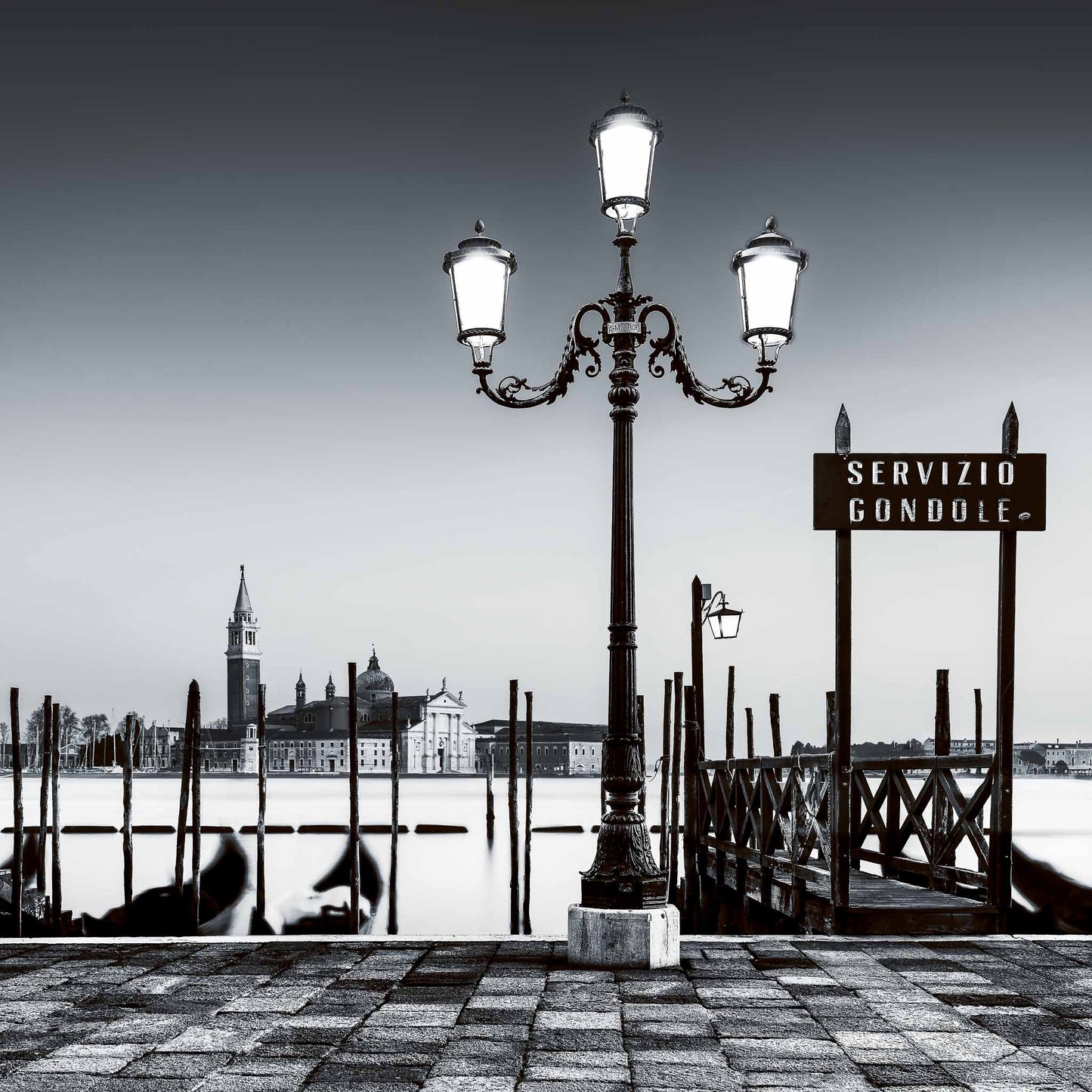 A fine art black and white photograph from Venice, Italy, showing a glowing lamp and gondola moorings at night with San Giorgio Maggiore beyond.