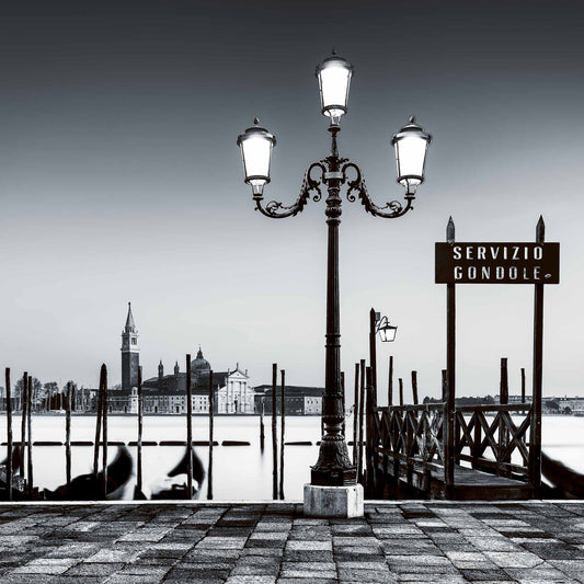 A fine art black and white photograph from Venice, Italy, showing a glowing lamp and gondola moorings at night with San Giorgio Maggiore beyond.