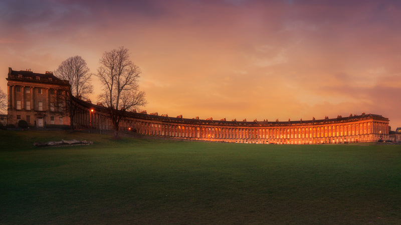 A fine art colour photograph of the Royal Crescent in Bath at sunset with warm light on the façade and a calm open lawn in the foreground.