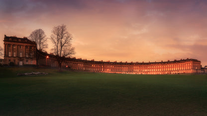 A fine art colour photograph of the Royal Crescent in Bath at sunset with warm light on the façade and a calm open lawn in the foreground.