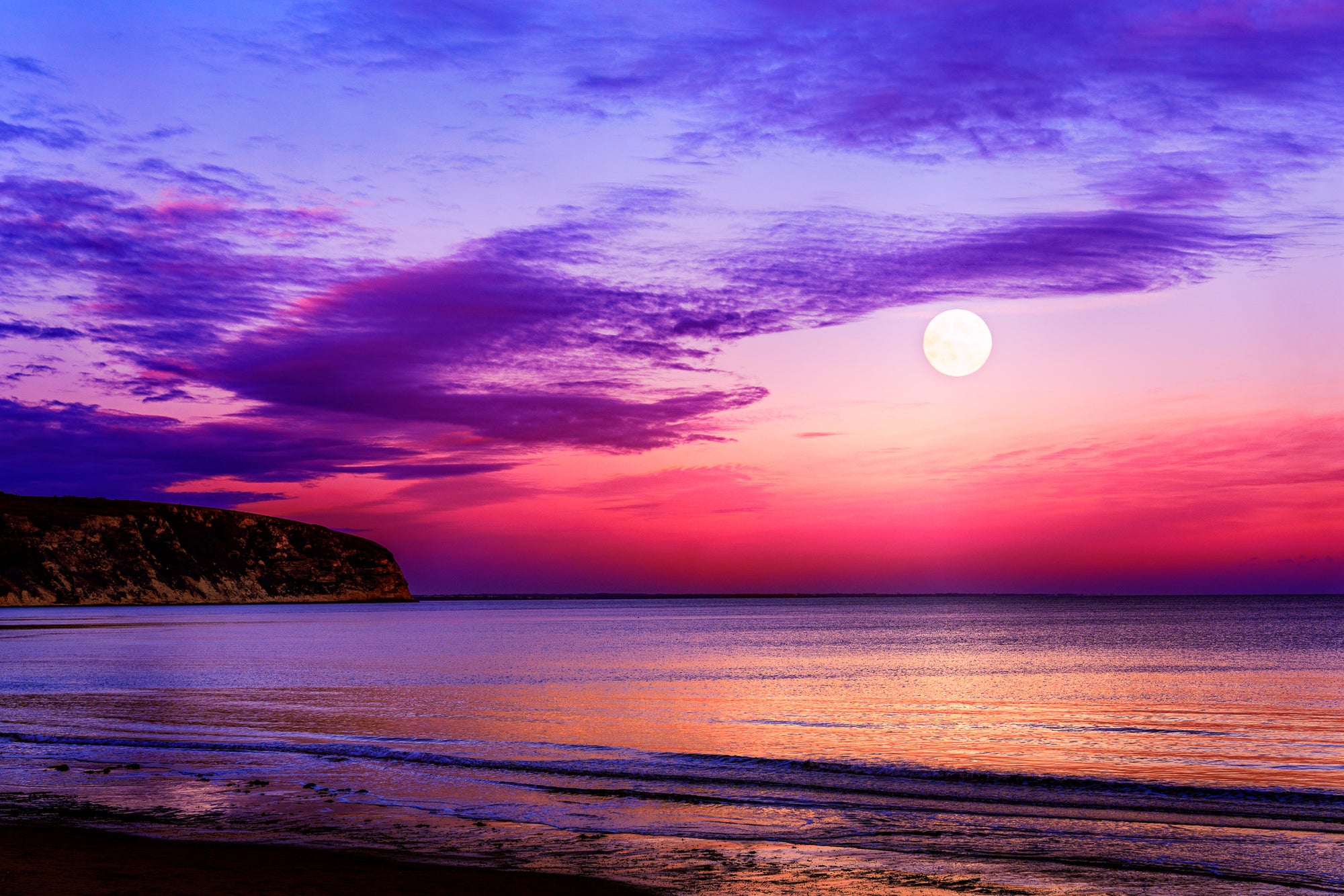 Moon rising over Swanage Bay at twilight with colourful sky reflecting on calm sea and gentle shoreline.