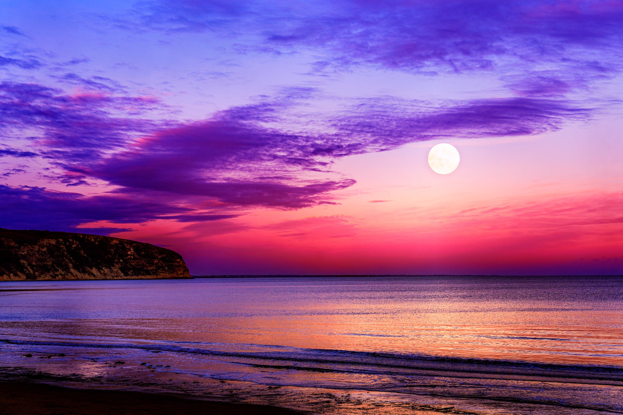 Moon rising over Swanage Bay at twilight with colourful sky reflecting on calm sea and gentle shoreline.