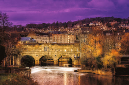 Pulteney Bridge in Bath at sunset with warm lights reflecting on the River Avon and historic buildings rising behind