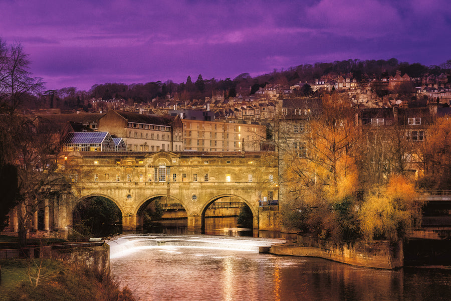 Pulteney Bridge in Bath at sunset with warm lights reflecting on the River Avon and historic buildings rising behind