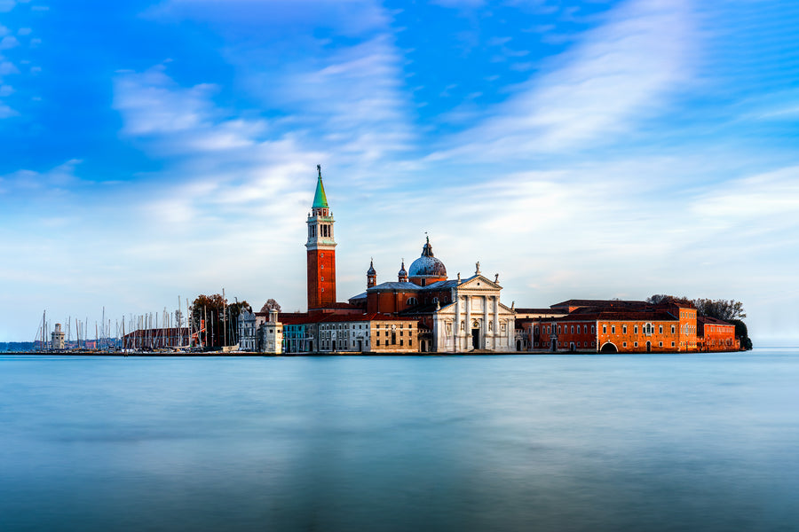 Colour photograph of San Giorgio Maggiore in Venice with church and bell tower reflected across calm lagoon water in daylight.