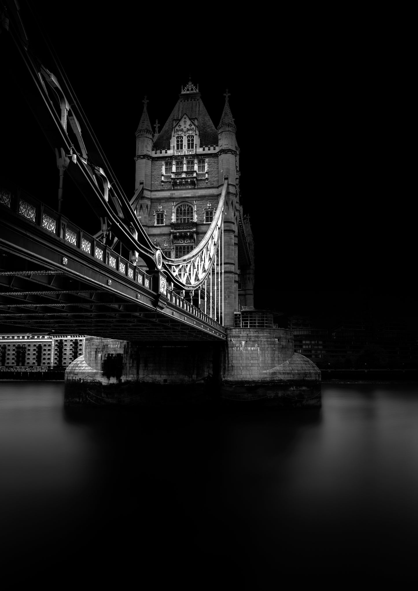 A fine art black and white photograph of Tower Bridge in London, viewed from beneath, showing its gothic structure and reflection on the Thames.