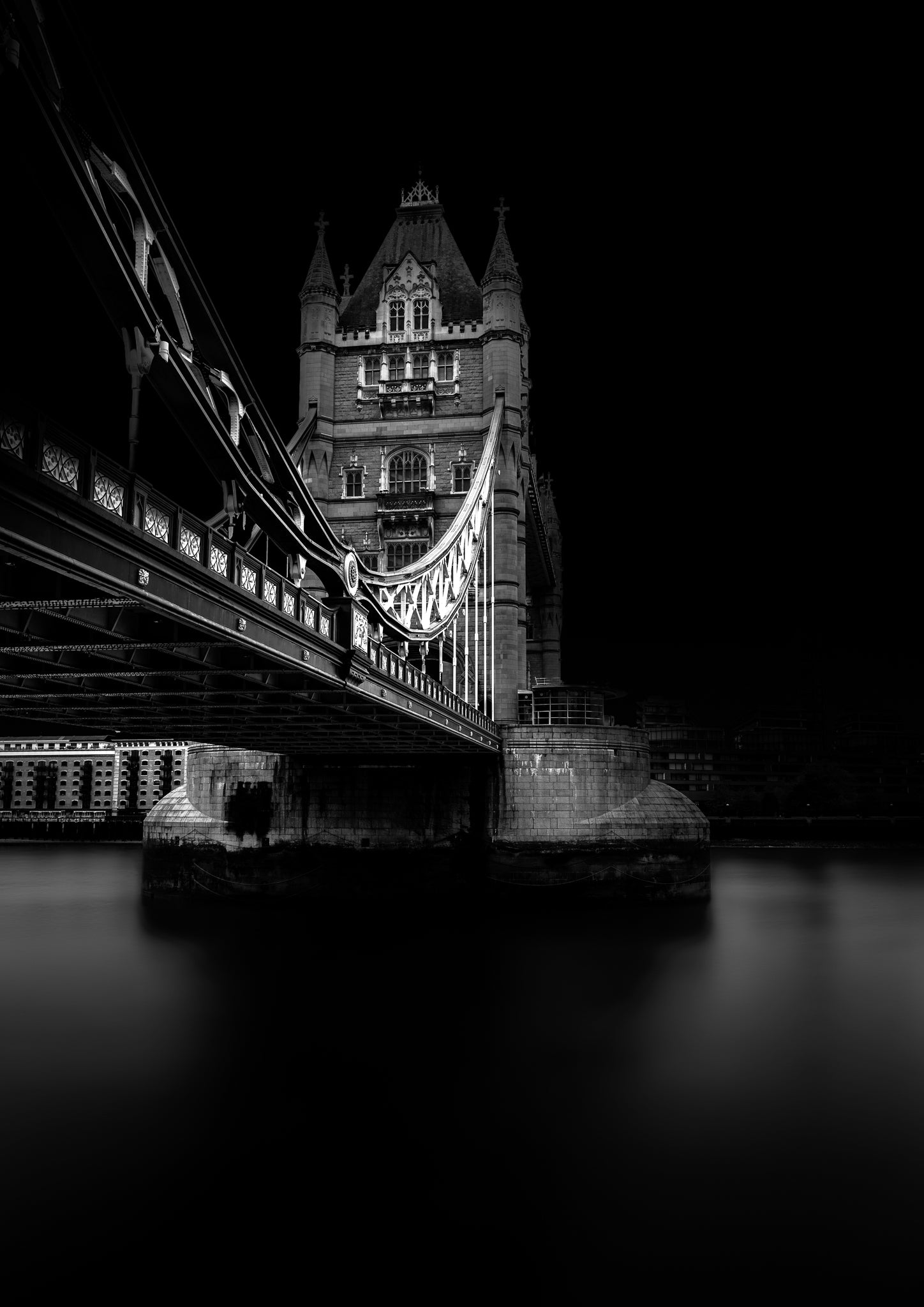 A fine art black and white photograph of Tower Bridge in London, viewed from beneath, showing its gothic structure and reflection on the Thames.