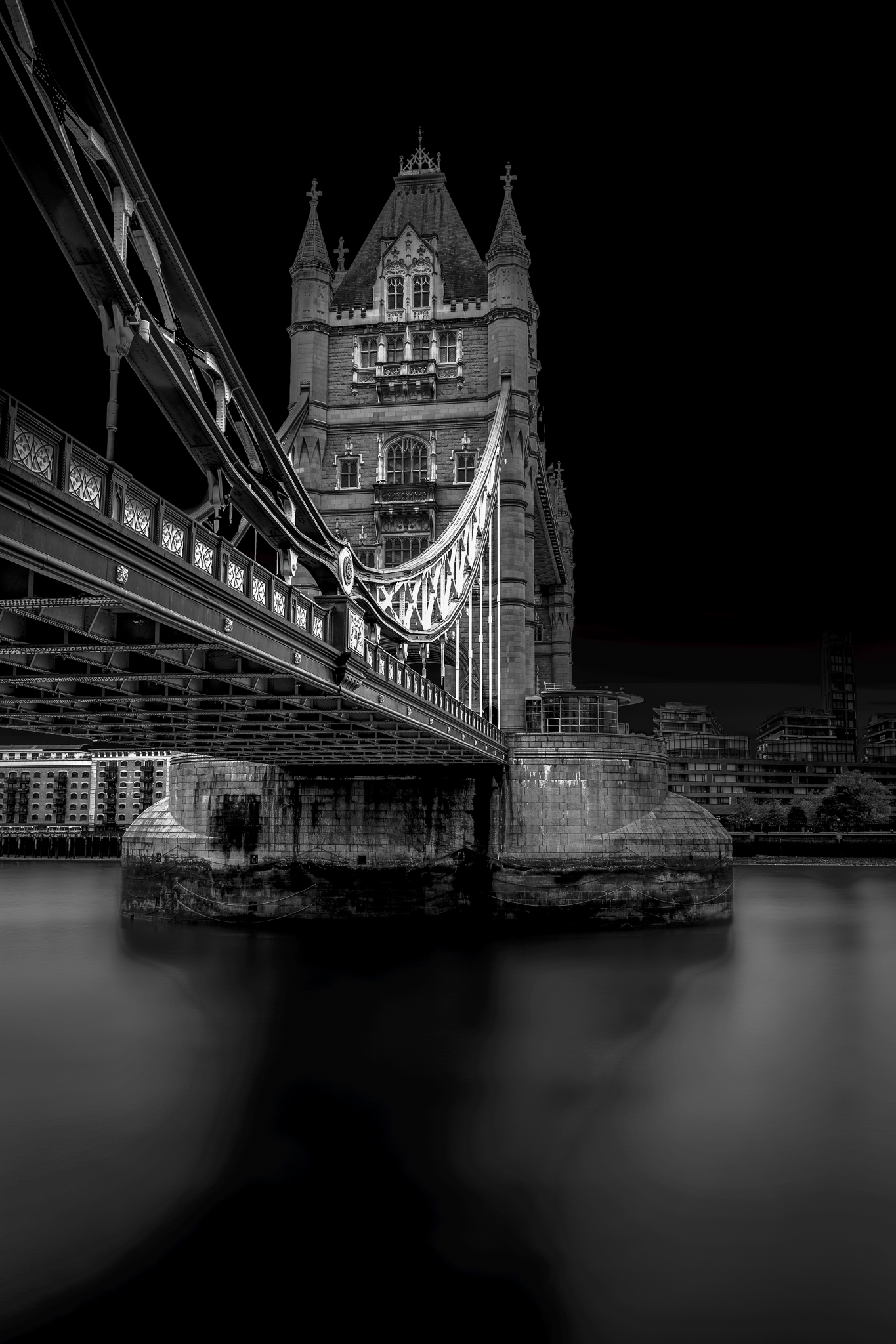 A fine art black and white photograph of Tower Bridge in London, viewed from beneath, showing its gothic structure and reflection on the Thames.