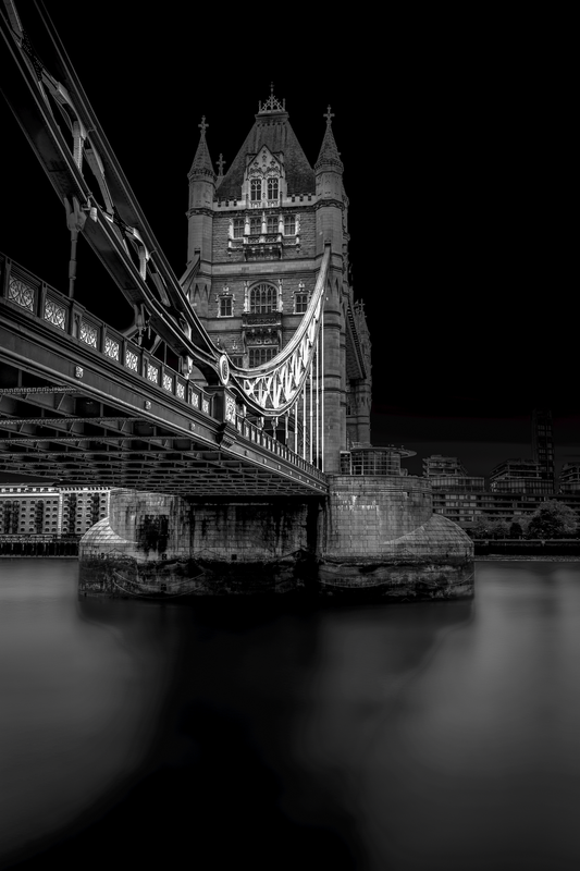 A fine art black and white photograph of Tower Bridge in London, viewed from beneath, showing its gothic structure and reflection on the Thames.