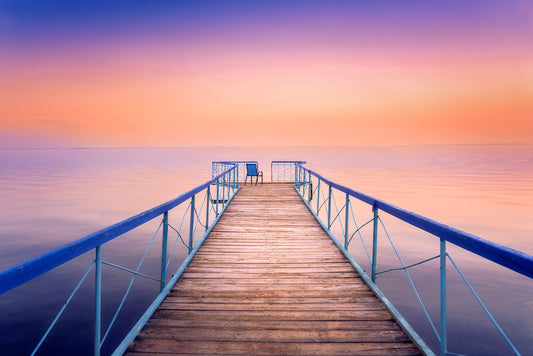 Fine art photograph of a wooden pier with a chair at sunset, overlooking calm water under a vibrant orange and purple sky.
