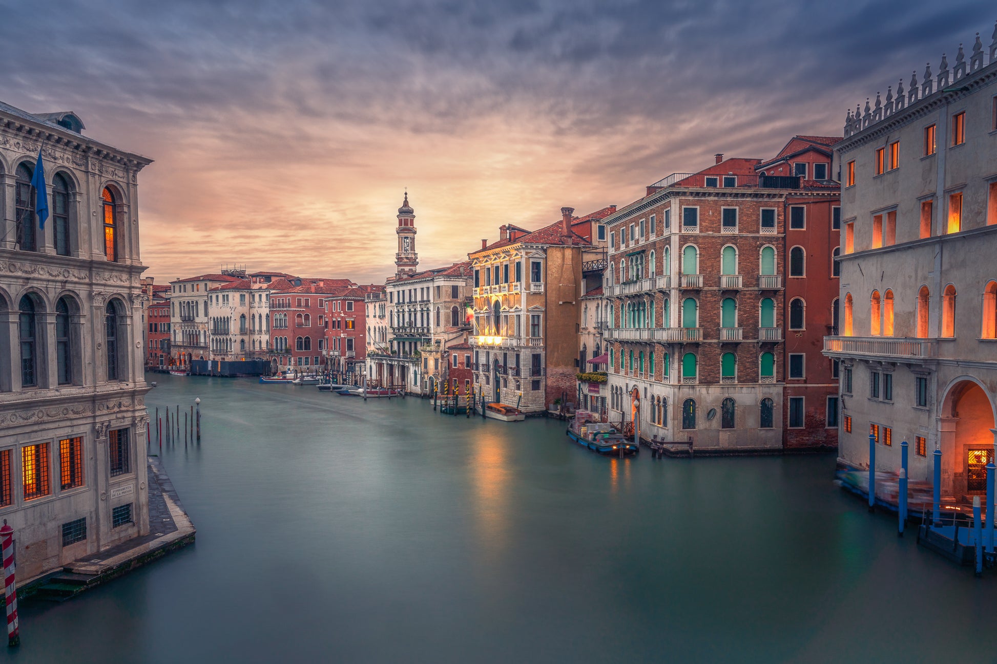 A fine art colour photograph of sunrise on the Grand Canal in Venice, with warm window light, calm long-exposure water, and historic buildings along the waterfront.