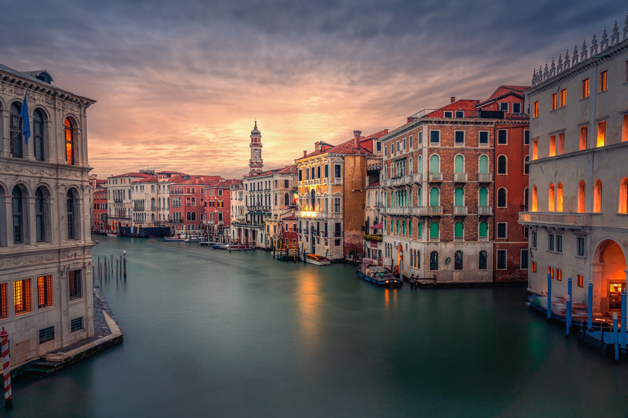 A fine art colour photograph of sunrise on the Grand Canal in Venice, with warm window light, calm long-exposure water, and historic buildings along the waterfront.