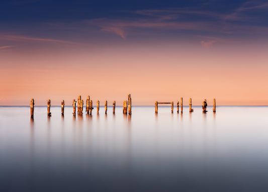Minimalist seascape of Swanage’s old pier, weathered wooden posts against soft peach and blue evening sky.