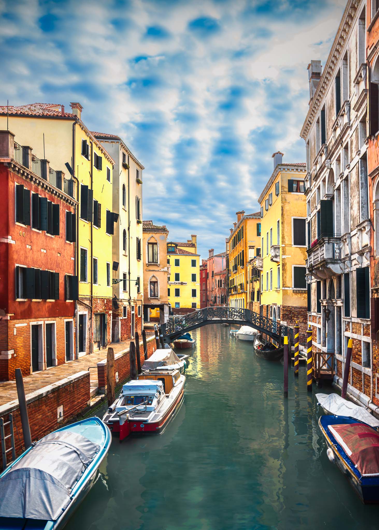 Fine art photograph of a Venetian canal with colorful facades, moored boats, and an iron bridge beneath a soft sky.