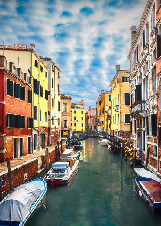Fine art photograph of a Venetian canal with colorful facades, moored boats, and an iron bridge beneath a soft sky.