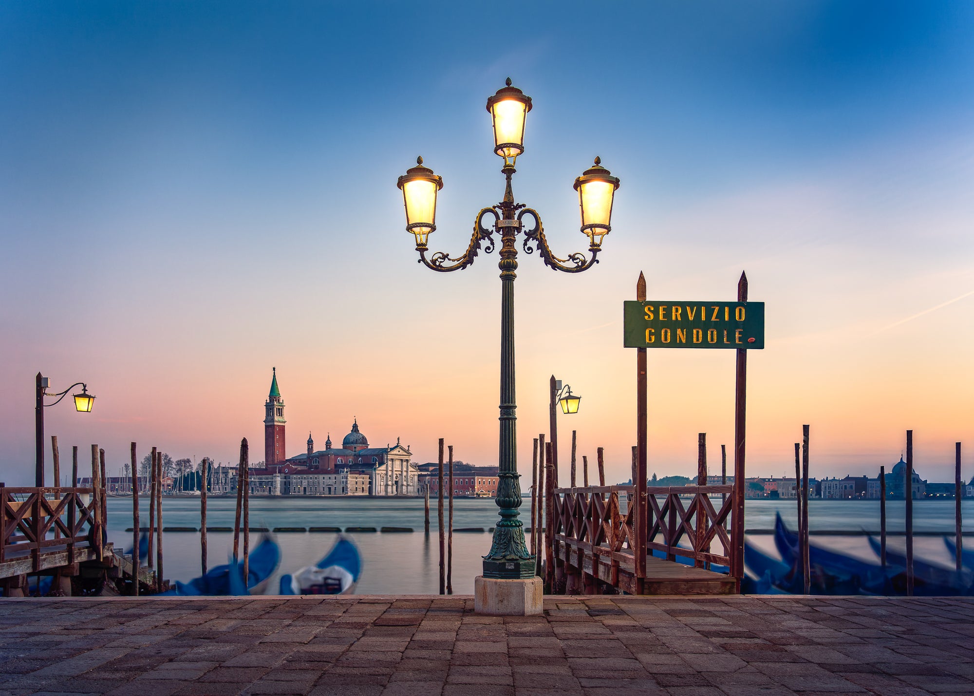 Sunrise on the Grand Canal in Venice with glowing street lamps, calm water, gondolas, and San Giorgio Maggiore in the background.